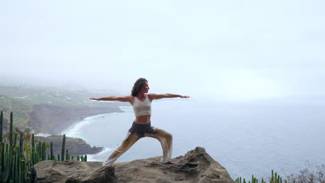 Balancing-in-the-yoga-warrior-pose-by-the-ocean,-beach,-and-rocky-mountains,-the-woman-embodies-motivation,-inspiration,-and-a-dedication-to-a-healthy-outdoor-lifestyle